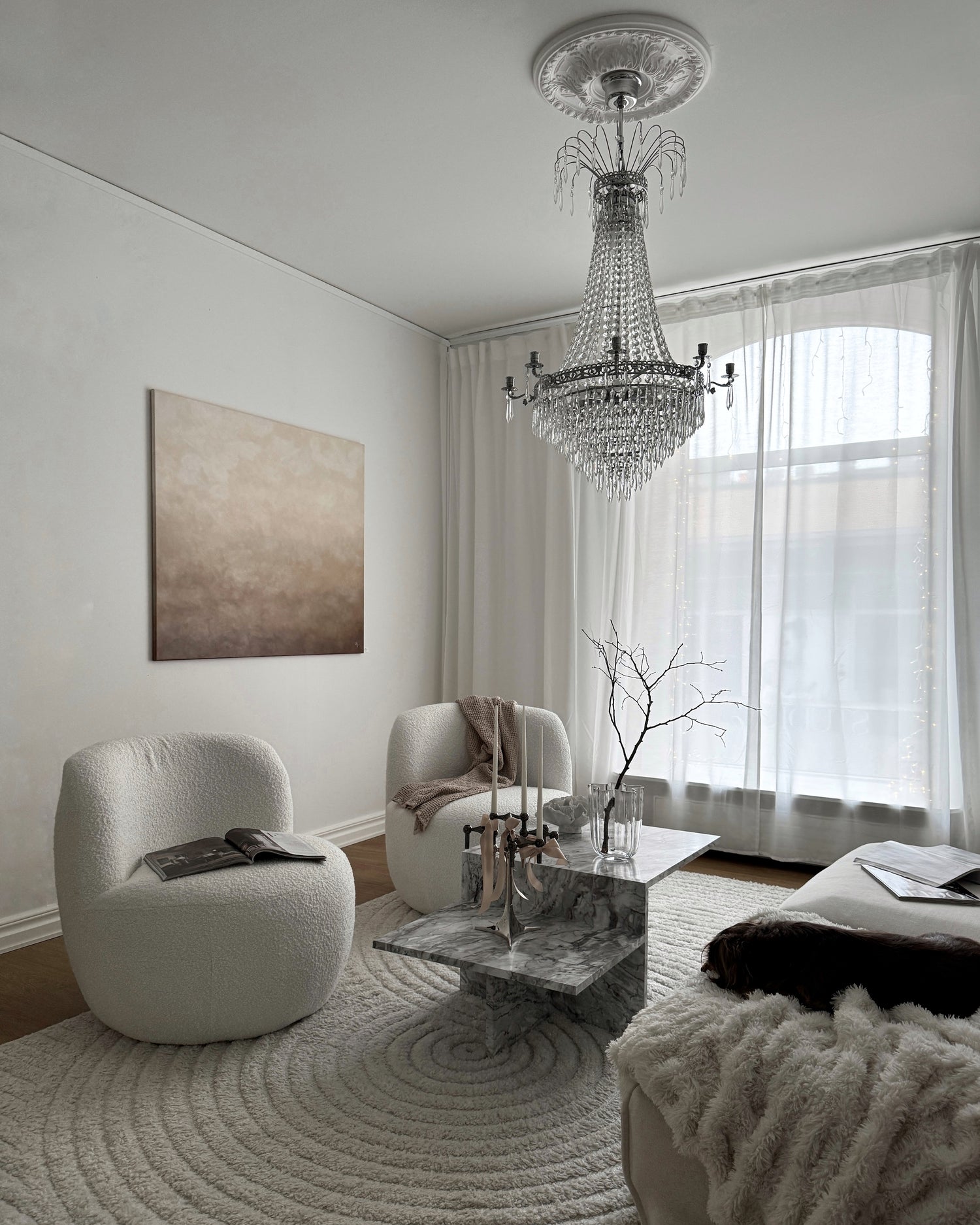 En bild på ett klassiskt inrett vardagsrum med kristallkrona, vita fåtöljer och en vacker handmålad tavla i brunt