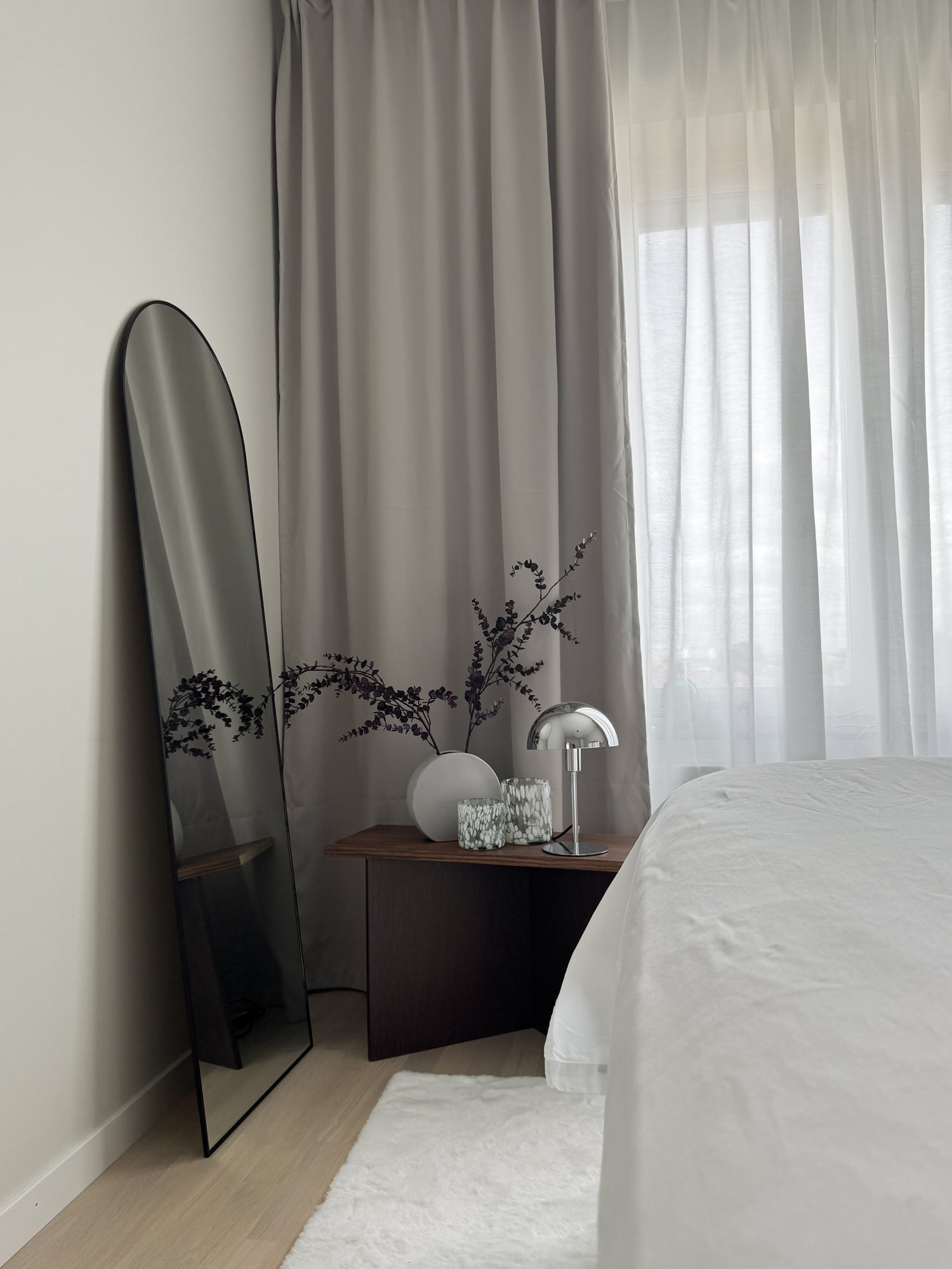 Ljust, mysigt sovrum i jordnära färger med toner av beige, vitt och mörkt trä. Ett litet sidobord med snygga detaljer och en spegel som står lutad mot väggen